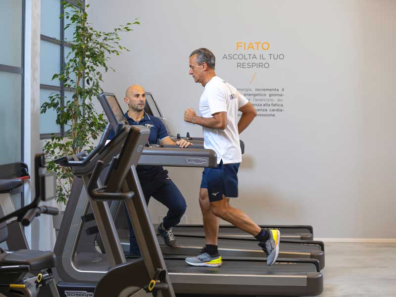 Hombre en el gimnasio corriendo suavemente en una cinta de correr para entrenamiento cardiovascular, seguido por un entrenador personal del Show Club que lo guía.