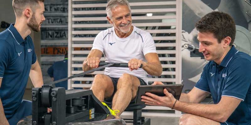 Hombre entrenando en el remo supervisado por sus entrenadores personales que le explican en la tablet los beneficios del entrenamiento de gimnasia suave